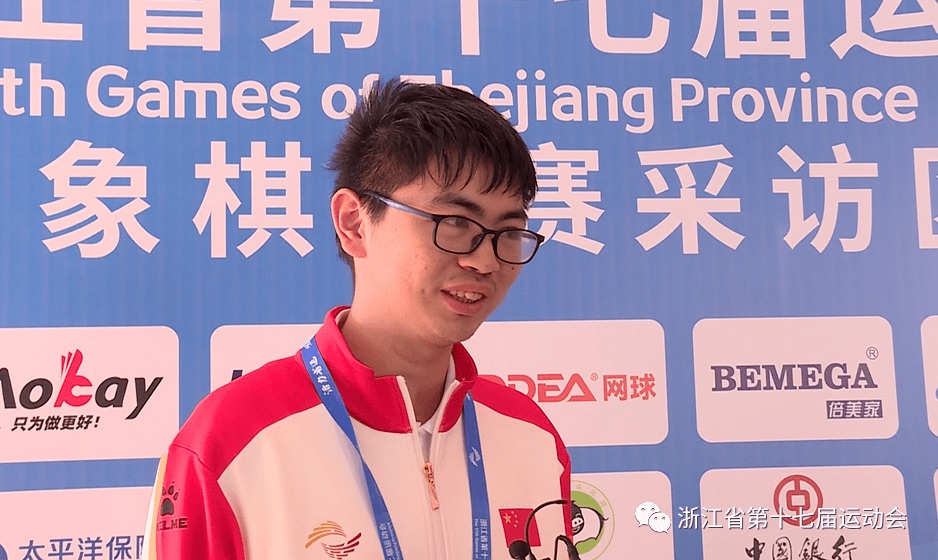 Yin Sheng, in a red and white Zhejiang team jacket and glasses, smiling at a sponsor backdrop after a game at the 17th Zhejiang Provincial Games in 2022.
